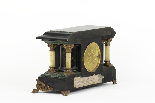 An angled reverse view of a Seth Thomas black mantle clock with green marbling detail. The Seth Thomas Clock Company logo is laid in a gold-toned door. On either side of the clock are white pillars with bronze-coloured base and top. The clock also features ornate gold claw feet and fine gold painted detailing. A torn maker's label is affixed to the back setting out the name 'Adamantine' and directions to use the clock.  