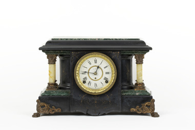 Obverse view of a Seth Thomas black mantle clock with green marbling detail. The clock face is white with black roman numerals and ornate gold rims. On either side of the clockface is a white pillar with bronze-coloured base and top. The clock also features ornate gold claw feet and fine gold painted detailing. 