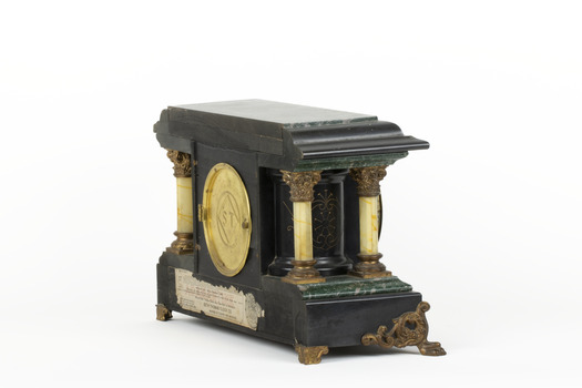 An angled reverse view of a Seth Thomas black mantle clock with green marbling detail. The Seth Thomas Clock Company logo is laid in a gold-toned back door. On either side of the clock are white pillars with bronze-coloured base and top. The clock also features ornate gold claw feet and fine gold painted detailing. A torn maker's label is affixed to the back setting out the name 'Adamantine' and directions to use the clock.  