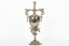 Elaborate metal trophy with a  woman in a flowing gown atop. The base features outward-curled feathers, while the handles are styled as birds. The trophy is also engraved with intricate filigree patterns.