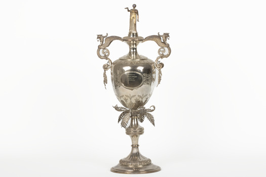 Elaborate metal trophy with a  woman in a flowing gown atop. The base features outward-curled feathers, while the handles are styled as birds. The trophy is also engraved with intricate filigree patterns.