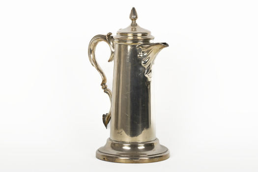 A golden coloured cupro-nickel electroplated hot water jug with a hinged lid, flat base, and elaborate handle.
