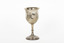 The side angle of a silver trophy. An engraved flower design is visible going around the side of the cup. On the left-hand side of the cup is a swirling deign with a small portion of writing visable.