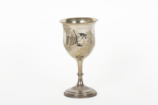 The side angle of a silver trophy. An engraved flower design is visible going around the side of the cup. On the left-hand side of the cup is a swirling deign with a small portion of writing visable.