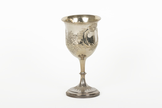The side angle of a silver trophy. An engraved flower design is visible going around the side of the cup. On the right-hand side of the cup is a swirling deign with a small portion of writing visible.