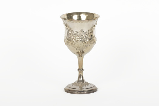 A silver trophy with an engraved flower design going around the cup. A seperate flower deign ring encircles the rim of the cup.