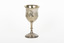 The side angle of a silver trophy. An engraved flower design is visible going around the side of the cup. On the left-hand side of the cup is a swirling deign with a small portion of writing visable.