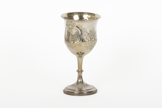 The side angle of a silver trophy. An engraved flower design is visible going around the side of the cup. On the left-hand side of the cup is a swirling deign with a small portion of writing visable.