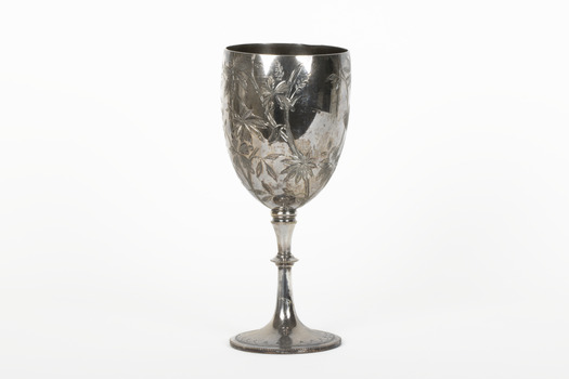 Silver cup in the shape of a wine glass, as the cup tapers to a pedestal. The cup is engraved with branches and leaves, the base engraved with lines, dots and a half-moo pattern. 