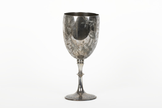 Silver cup in the shape of a wine glass, as the cup tapers to a pedestal. The cup is engraved with branches and leaves, the base engraved with lines, dots and a half-moo pattern. 
