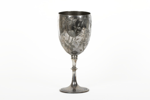 Silver cup in the shape of a wine glass, as the cup tapers to a pedestal. The cup is engraved with branches and leaves, the base engraved with lines, dots and a half-moo pattern. 