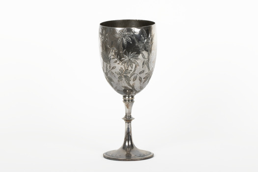 Silver cup in the shape of a wine glass, as the cup tapers to a pedestal. The cup is engraved with branches and leaves, the base engraved with lines, dots and a half-moo pattern. 