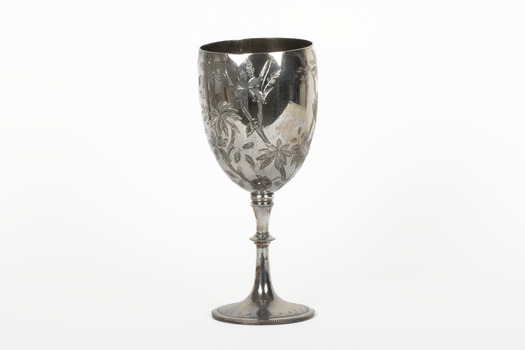 Silver cup in the shape of a wine glass, as the cup tapers to a pedestal. The cup is engraved with branches and leaves, the base engraved with lines, dots and a half-moo pattern. 