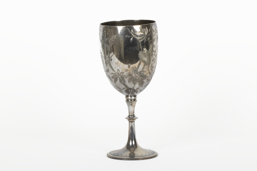 Silver cup in the shape of a wine glass, as the cup tapers to a pedestal. The cup is engraved with branches and leaves, the base engraved with lines, dots and a half-moo pattern. 