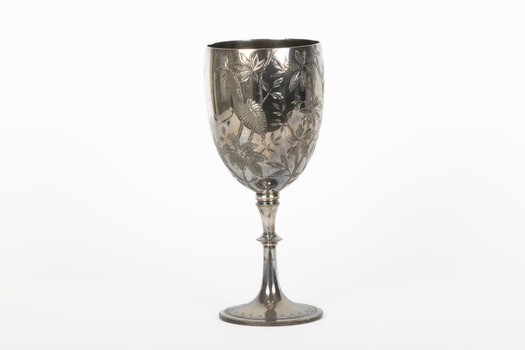 Silver cup in the shape of a wine glass, as the cup tapers to a pedestal. The cup is engraved with branches and leaves, the base engraved with lines, dots and a half-moo pattern. 