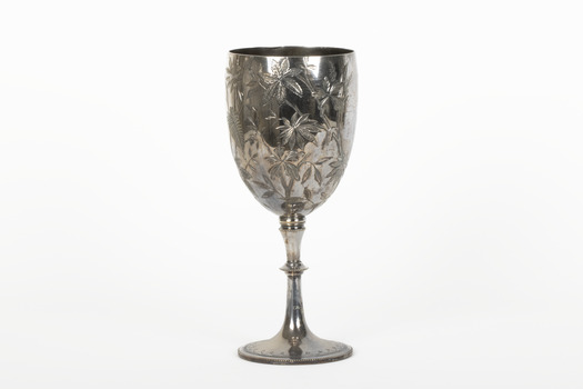 Silver cup in the shape of a wine glass, as the cup tapers to a pedestal. The cup is engraved with branches and leaves, the base engraved with lines, dots and a half-moo pattern. 