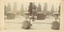 A stereoscopic photograph mounted on card depicting Town Hall Gardens covered in snow. A fountain is at the centre, with a fluted garden urn and a tap stand in the foreground. A row of trees line the background with a church steeple visible in the distance.  
