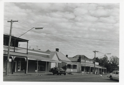 Black and white photograph of a street lined with shops, one of which is a butcher. Two parked cars are along the road, one on the left side of the road and one on the right.