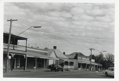 Black and white photograph of a street lined with shops, one of which is a butcher. Two parked cars are along the road, one on the left side of the road and one on the right.