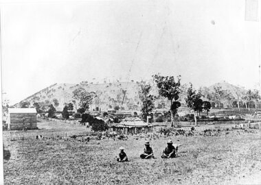 Three First Nations people seated in front of a small volcanic hills; scattered buildings, fences and trees in background.