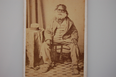 Bearded Aboriginal man in European dress sitting on a wooden chair beside a small table with a hat on top.