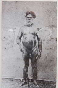 Naked Aboriginal man standing with walking stick in hand.