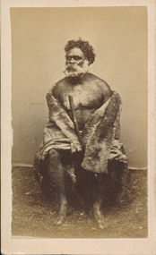 Aboriginal man sitting in traditional animal skin cloak, holding a walking stick.