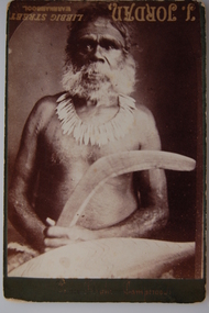 Bearded, tattooed Aboriginal man wearing a kangaroo tooth necklace, holding a boomerang and shield
