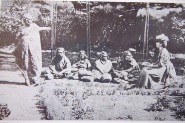 Informal gathering of five First Nations people in traditional dress, in conversation with a seated European lady with notebook in hand.