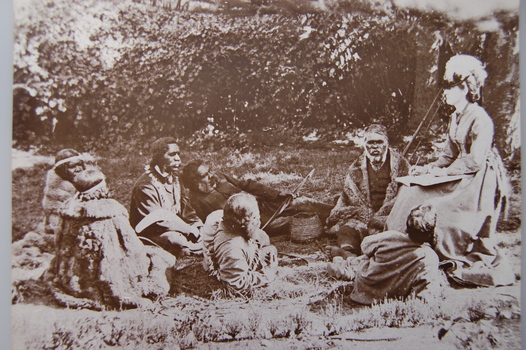 Informal gathering of seven First Nations people in traditional dress, in conversation with a seated European lady with pen and notebook in hand.  