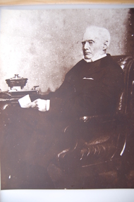 Elderly bearded man wearing Chesterfield overcoat sitting on a upholstered wooden chair, legs crossed, holding a piece of paper.