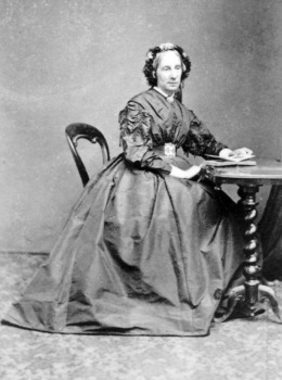 Middle aged Victorian lady sitting at writing table wearing Victorian era gown and headwear