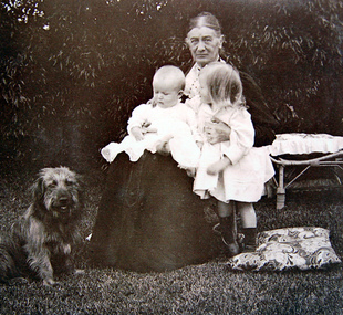 Elderly lady seated in garden holding two young children, with a dog beside her.