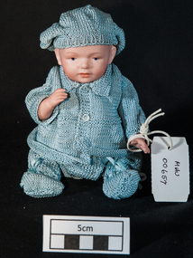 A very small celluloid doll dressed as a boy in fine hand -knitted hat, vest, jacket and pants.