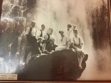 A party of 8 people grouped on a large rock in front of a waterfall.  3 men, standing, the  first a cigarette in his mouth and holding a hat; the second holds a hiking pole; 3 women, the third has her elbow raised with her hand on the back of her head; and 2 youths, the bigger wears braces and sits at the front of the rock, feet crossed, hands in his lap.   The Rock has letters B.P  and t E (?) painted on it. 
