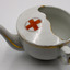 White china cup with teapot-like spout and half-enclosed opening on top and small handle on the side adjacent to spout. Gold edge painted detail and red cross on top.