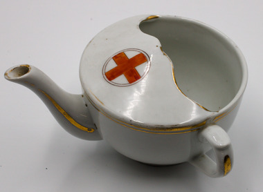 White china cup with teapot-like spout and half-enclosed opening on top and small handle on the side adjacent to spout. Gold edge painted detail and red cross on top.