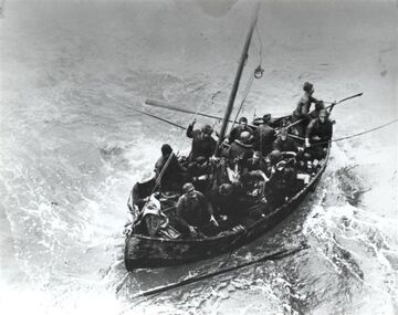 lifeboat at sea with approximately 16 crew members of the sunken Peter Silvester aboard. Some are standing but most are sitting. One of the oars has fallen into the water but there is a line that has been thrown to the boat to facilitate the rescue.