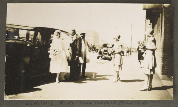 Sepia toned photograph of bride and attendants in the courtyard of the Mission to Seafarers. The bride's dress is calf length and she has just stepped out of a large car assisted by her Father Mr Walker. The Bride is holding a large bouquet. There are two attendants or Bridesmaids to the right of the photograph, wearing  layered calf length dresses, cloche hats  and holding a small bouquet. Both are standing outside the Mission to Seamen  on Flinders St waiting to assist the bride. 