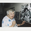 Colour photograph of Edith Warburton looking at a large black and white picture of Padre Oliver and seamen in the 50s.