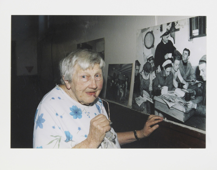 Colour photograph of Edith Warburton looking at a large black and white picture of Padre Oliver and seamen in the 50s.