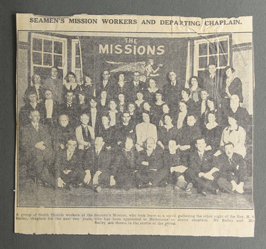 Newspaper clipping with a black and white photograph depciting a large group of people sitting and standing in front of the Mission's flag.