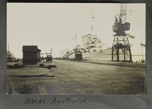 Small landscape sepia image depicting 3-funnel stack battleship moored at a pier, an ships ensign flies at far right of frame.