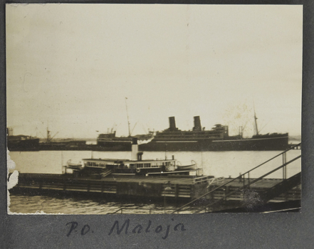 small landscape sepia photograph adhered to an album page. Depicts 2 vessels  an ocean going 2 stack ship in the distance and a smaller vessel (ferry or coaster) moored against a pier.
