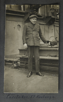A sepia image of an older gentleman with moustache in a long jacket and a flat cap stands on a ship deck near a cargo hatch. 