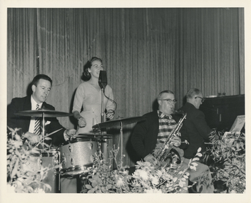 Small black and white photograph depicting a band playing on stage in the Mission's hall: from left to right three male musicians sitting down: a drummer, a saxophonist and the pianist, behind the saxophonist is standing a female singer singing with a microphone on a stand. Behind the band the curtains are drawn, in front of them the stage is decorated with 3 vases of large flowers.