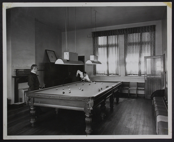 Chaplains playing pool in the Billiard room in Flinders Street Mission, one is with the cue above the table, the other is waiting standing to the fireplace.