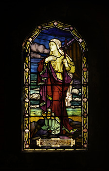 Arched stained glass window depicting Mary with a cross, in the background the sail of a small barque on the sea with waves and sky