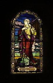 Arched stained glass window depicting Mary with a cross, in the background the sail of a small barque on the sea with waves and sky