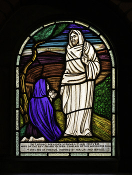 Stained glass window depicting Mary in the traditional blue cloth kneeling down in front of Jesus standing, wearing a white cloth. 